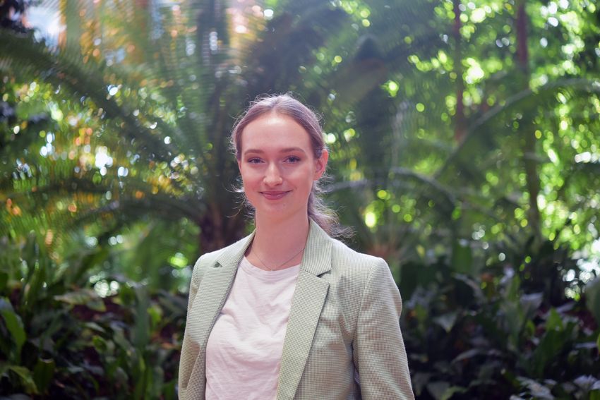 Bettina Dietmair is smiling, wearing a white top and green jacket. She is standing in front of a leafy backdrop.