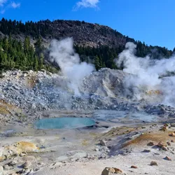 A photograph of a geothermal lake in Lassen Volcanic National Park. There is a raised mountain in the background covered in rocks with tall trees further down. In the foreground, the ground is rocky, with a blue lake in the middle of the image that has steam coming off of it. 