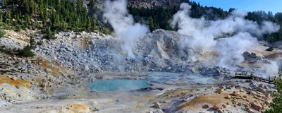 A photograph of a geothermal lake in Lassen Volcanic National Park. There is a raised mountain in the background covered in rocks with tall trees further down. In the foreground, the ground is rocky, with a blue lake in the middle of the image that has steam coming off of it. 