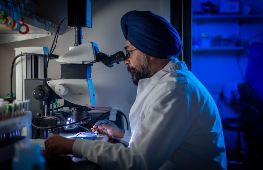 Harmit Malik is in the lab, wearing a white coat and studying something under a microscope. Behind him is a glowing blue background. Harmit Malik is in the lab, wearing a white coat and studying something under a microscope. Behind him is a glowing blue background.