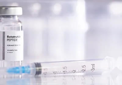 A glass vial of peptide drug retatrutide with a syringe for injection, against a white backdrop. 