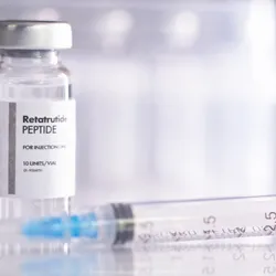 A glass vial of peptide drug retatrutide with a syringe for injection, against a white backdrop. 