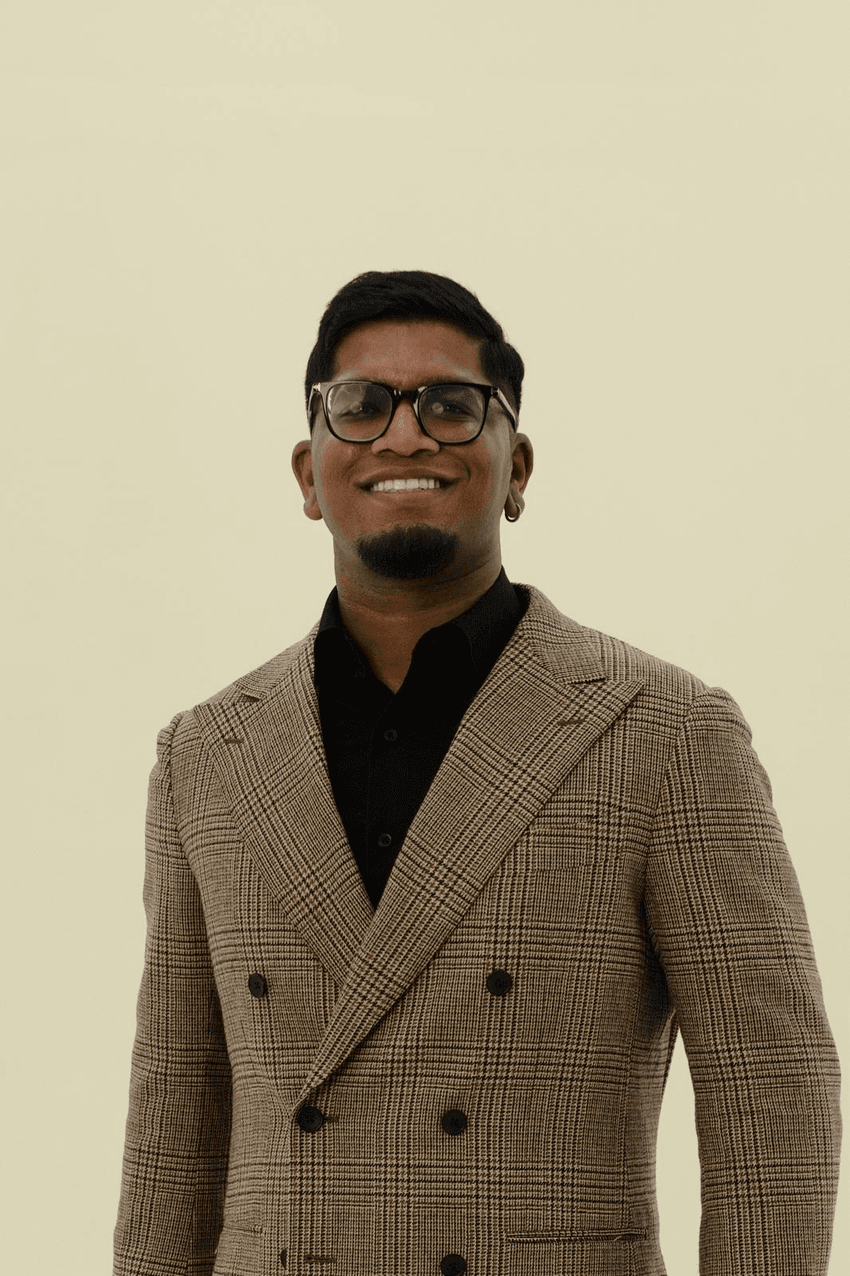 Karthik Achari smiles against a cream backdrop, wearing black-framed glasses, a black shirt, and a double-breasted jacket with a brown plaid pattern. 