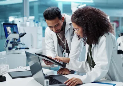 Two people, one a clinical trial monitor, discuss clinical trial needs in a laboratory setting.