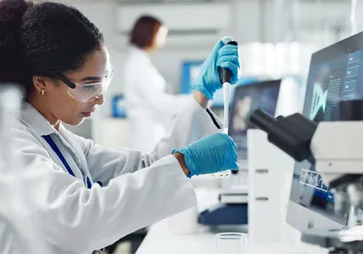 A scientist pipetting a small-volume biological sample in an analytical laboratory, with mass spectrometry data visible on a monitor, illustrating biopharmaceutical characterization workflows for AAV analytics and viral vector quantification.
