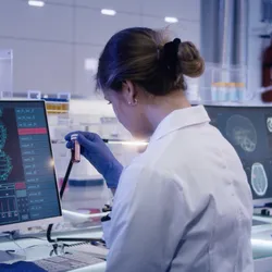 Image of two scientists sitting side by side in a laboratory. The woman on the left is looking at a vial while a man on the right is looking through a microscope.