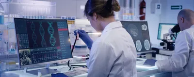 Image of two scientists sitting side by side in a laboratory. The woman on the left is looking at a vial while a man on the right is looking through a microscope.
